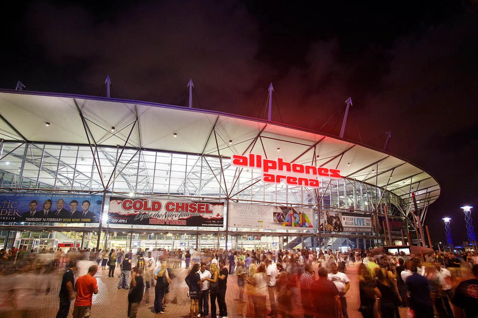 The curved roof and glass front of the arena at Sydney Olympic Park lit up at night with a crowd outside